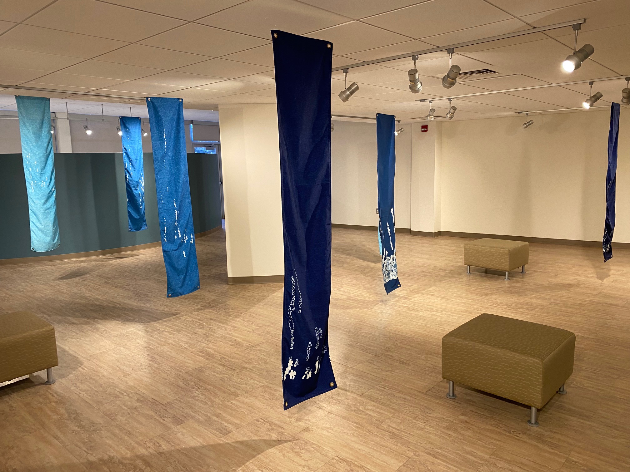 A view of Liz Miller's exhibit in the Regier Art Gallery: long vertical strips of fabric in various shades of blue with representations of the sounds in white