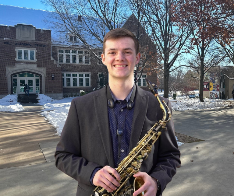 Eli Regier in a brown suit jacket and dark blue shirt, holding his saxophone, standing on the Bethel campus with Will Academic Center in the background