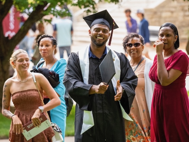 A graduate in a black robe with a light-colored stole and mortarboard hat, holding his diploma, celebrates with family.