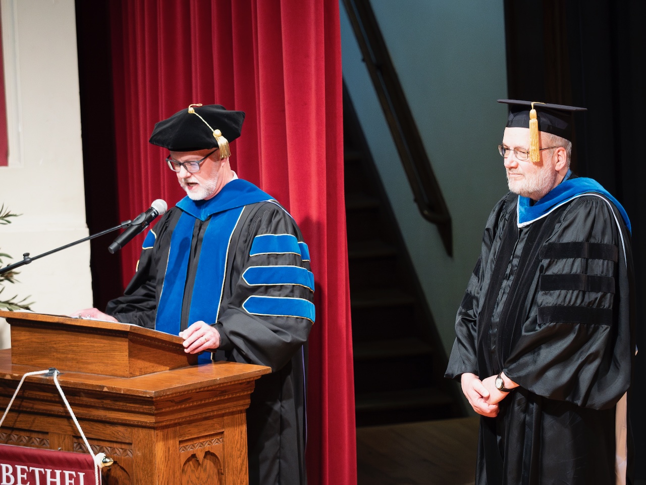 Bob Milliman reads the Schrag Teaching Award citation on the Mem Hall podium as Mark Jantzen looks on, both dressed in academic regalia