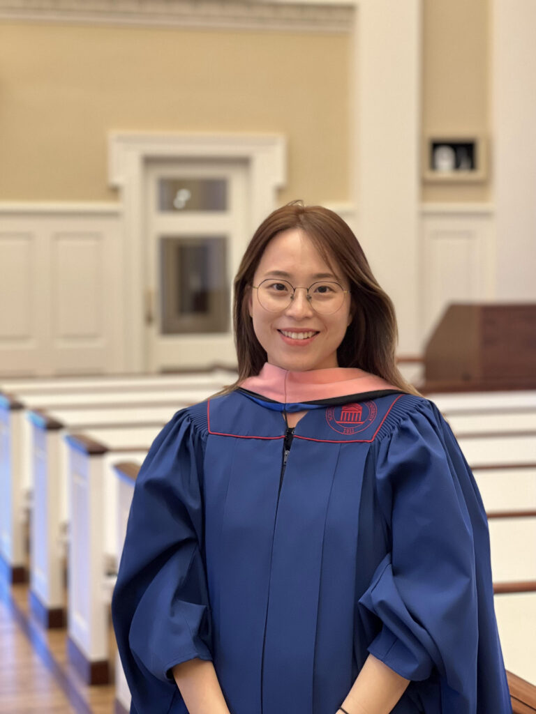 YunJung Lee in organist's blue robe with stole, standing in a church with pews behind her