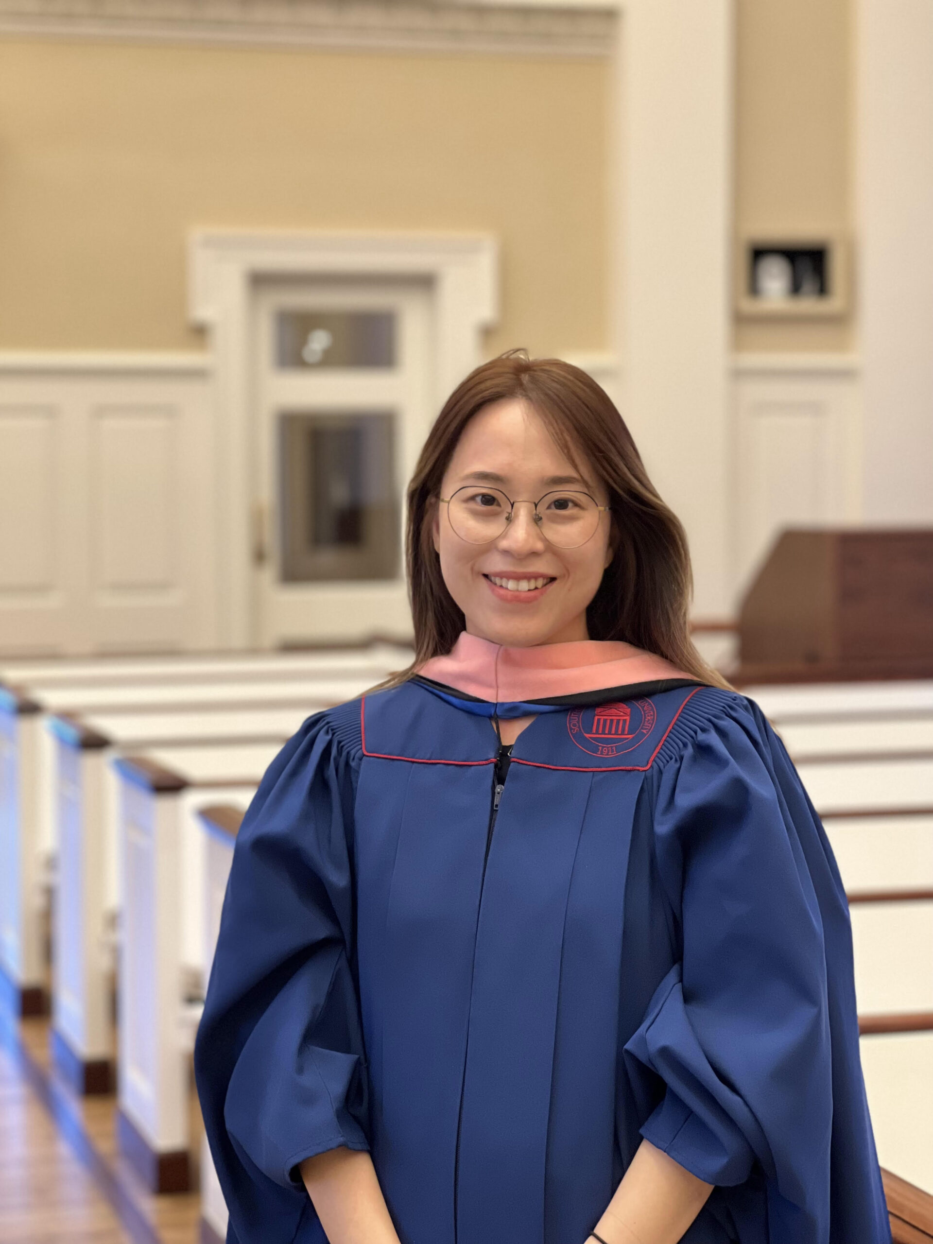 YunJung Lee in organist's blue robe with stole, standing in a church with pews behind her
