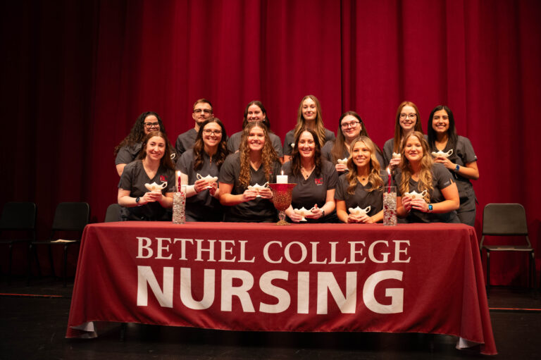 Group photo of the nursing Class of 2027, standing behind a table with a Bethel College Nursing cloth, and holding their lamps