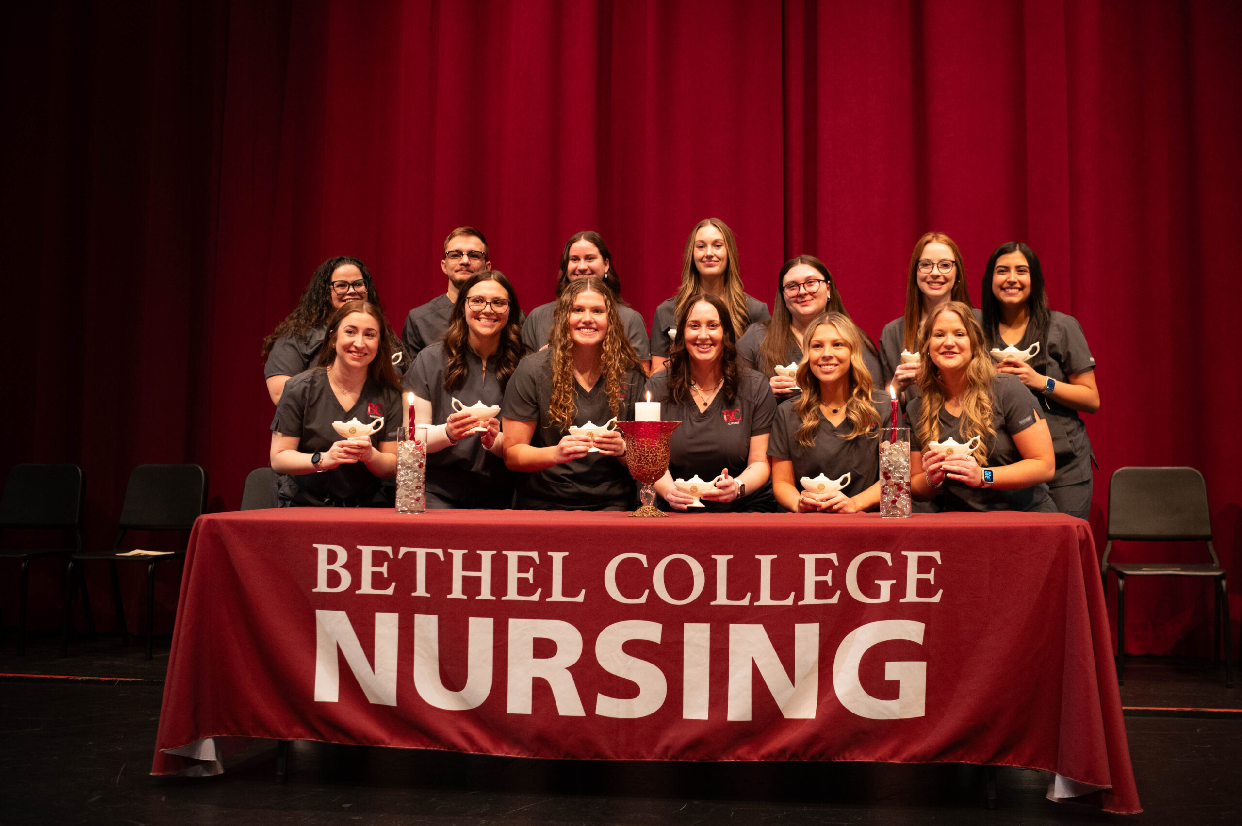 Group photo of the nursing Class of 2027, standing behind a table with a Bethel College Nursing cloth, and holding their lamps
