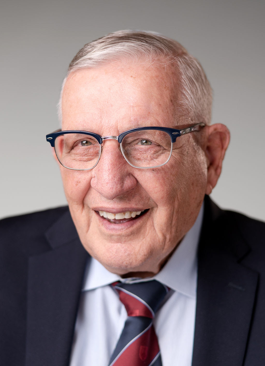 White man with gray hair, wearing glasses and a suit coat, white shirt and red-and-navy tie