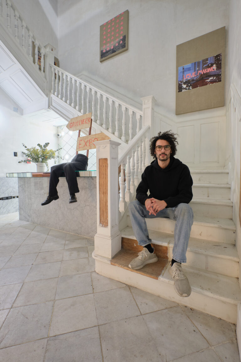 Rodrigo Cacho, a young Latinx man with dark curly hair wearing a black shirt, gray pants and glasses, sitting at the bottom of a long white staircase.