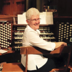 White woman with short curly white hair, wearing a white jacket and looking over her shoulder while sitting at the console of a large pipe organ